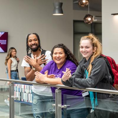 Three female students and one male student with dreadlocks pose with the Riverbat sign against a metal and glass rail at ACC Highland campus.